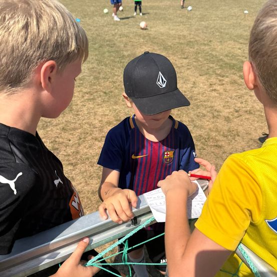 Drei Jungen stehen in der Nähe eines Fußballtors, einer hält eine Clipboard und einen Stift, wahrscheinlich zur Punktenotierung. Der Junge in der Mitte trägt eine schwarze Kappe mit einem weißen Logo. Im Hintergrund spielen andere Kinder Fußball.