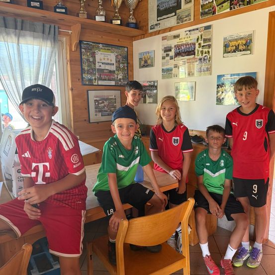 Eine Gruppe junger Fußballspieler in verschiedenen Teamtrikots posiert für ein Foto in einem Raum mit Trophäen und Bildern an der Wand.