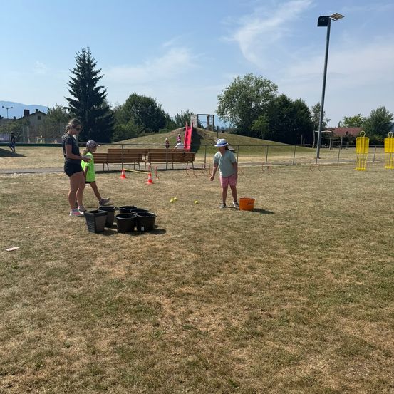 Zwei Frauen und ein Kind spielen ein Spiel mit Eimern und Bällen auf einem Rasenfeld. Sie stehen vor einer Reihe von Eimern. In der Ferne befindet sich ein Spielplatz mit einer Rutsche und einer Bank.