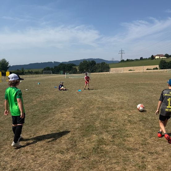 Eine Gruppe von Kindern spielt Fußball auf einem Grasfeld mit klarem Himmel und Bergen im Hintergrund.