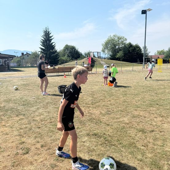 Ein junger Junge übt Fußball auf einem Feld mit anderen Kindern und Erwachsenen. Bäume und ein Gebäude sind im Hintergrund.
