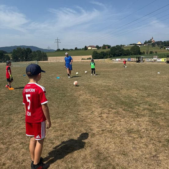 Ein Fußballtraining mit Kindern in roten und grünen Trikots, einem Trainer in Blau und einer hügeligen Landschaft im Hintergrund.