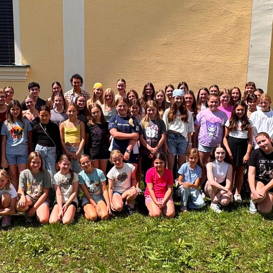 Eine Gruppe junger Menschen, möglicherweise Schüler, posiert für ein Foto vor einem Gebäude mit gelber Wand. Einige stehen, andere sitzen auf dem Rasen.