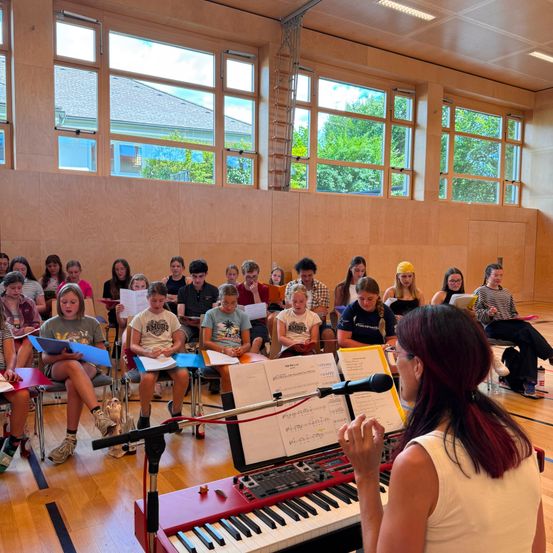 Eine Gruppe von Schülern in einem Klassenzimmer, die um ein Klavier herumsitzen, mit einer Frau vor ihnen, die in ein Mikrofon singt.