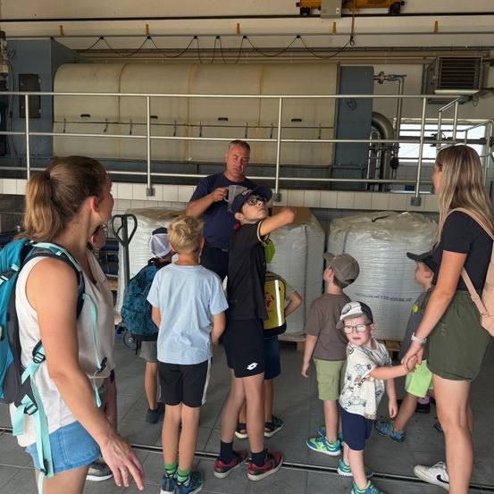 Eine Gruppe von Kindern und Erwachsenen versammelt sich in einer Fabrikumgebung, wobei ein Mann in einem blauen Shirt ihnen etwas erklärt. Die Kinder tragen Hüte und Brillen, und im Hintergrund befinden sich große weiße Säcke.