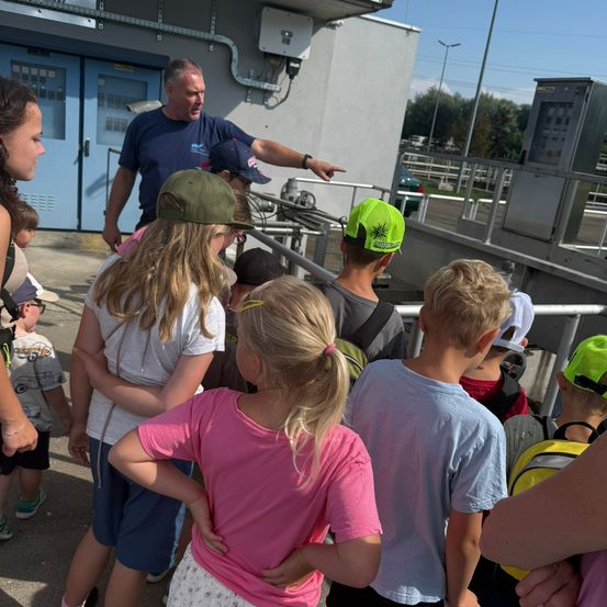 Eine Gruppe von Kindern und einem Mann in einem blauen T-Shirt versammelt sich vor einem Gebäude, möglicherweise für eine Tour. Der Mann zeigt auf etwas, während die Kinder aufmerksam zuhören.