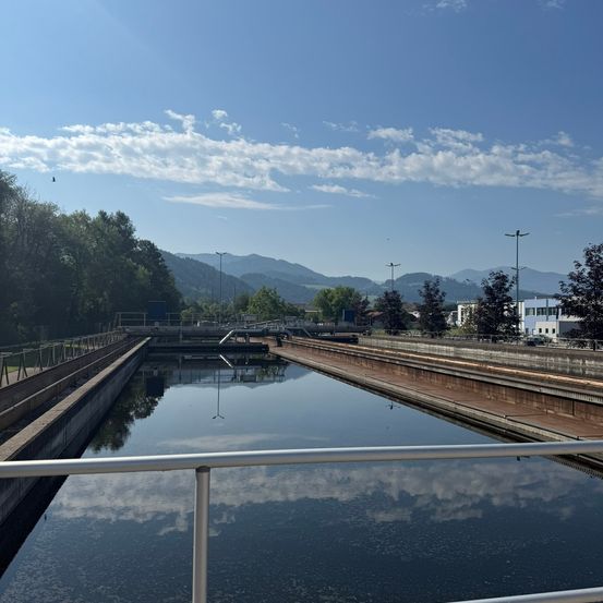 Ein ruhiger Blick auf einen Wasserkanal mit klarem blauem Himmel, Bergen in der Ferne und Bäumen an den Seiten.
