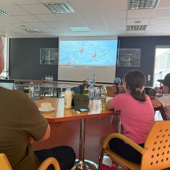 Eine Gruppe von Menschen sitzt um einen Tisch in einem Raum und schaut sich eine Präsentation auf einer Projektionsleinwand an. Flaschen, Tassen und eine Kappe sind auf dem Tisch. Der Raum hat eine Projektionsleinwand, Deckenleuchten und eine Tür.