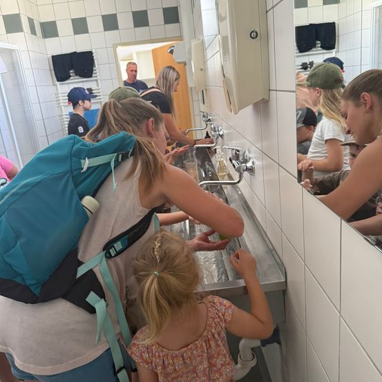 Eine Frau und ein junges Mädchen waschen ihre Hände in einem Waschbecken in einem Badezimmer. Die Frau trägt einen Rucksack.