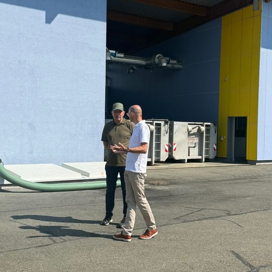 Zwei Männer stehen vor einem Gebäude mit einer blau-gelben Wand. Ein Mann trägt einen Hut und der andere ein weißes Hemd. Sie führen ein Gespräch.