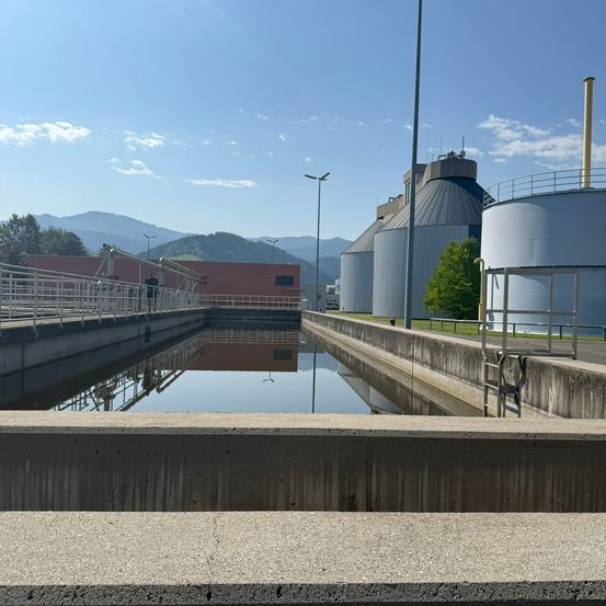 Eine Wasseraufbereitungsanlage mit großen Tanks und einer Brücke über einem Kanal, unter einem teilweise bewölkten Himmel.