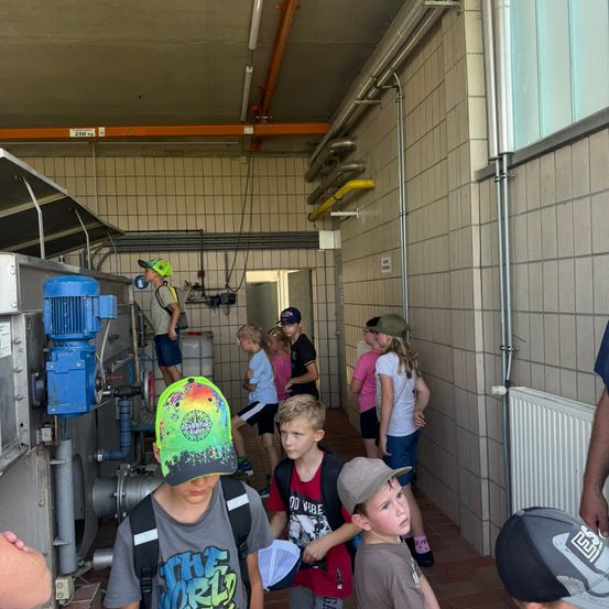 Eine Gruppe von Kindern und einem Erwachsenen stehen in einem Flur mit Industriegeräten. Die Kinder tragen Hüte und Rucksäcke. Der Erwachsene steht neben einer blauen Maschine.