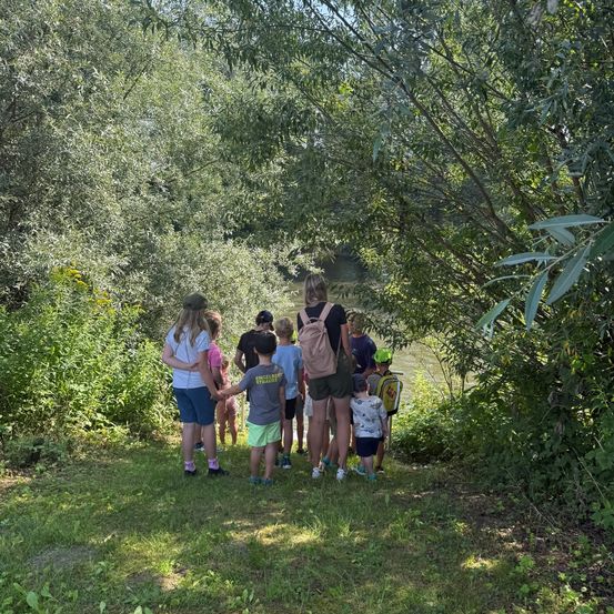 Eine Gruppe von Kindern und Erwachsenen steht in der Nähe eines Flusses, möglicherweise auf einem Naturspaziergang. Einige tragen Hüte und Rucksäcke.