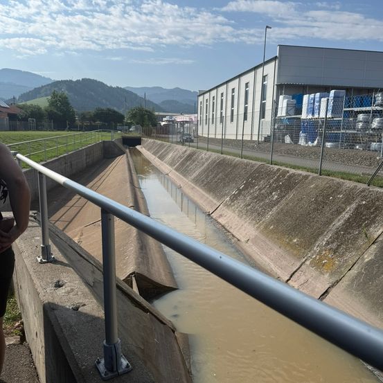 Ein Kanal mit fließendem Wasser, begrenzt von einer Betonmauer, mit einer Person auf der linken Seite. Im Hintergrund befindet sich ein Gebäude mit Zaun und einer Bergkette.
