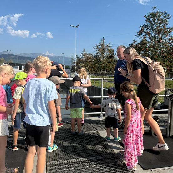 Eine Gruppe von Kindern und Erwachsenen steht auf einer Metallgitterplatte, möglicherweise in einer Wasseraufbereitungsanlage. Sie tragen Freizeitkleidung und einige tragen Hüte. Eine Frau in der Mitte hält die Hand eines Kindes. Hinter ihnen befindet sich ein Metallgeländer, Bäume und eine Straßenlaterne. In der Ferne gibt es Berge und einen klaren blauen Himmel.