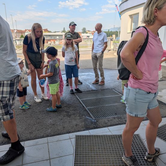 Eine Gruppe von Menschen, einschließlich Erwachsener und Kindern, steht auf einer nassen Betonfläche mit einem Metallrost. Sie warten wahrscheinlich auf etwas.