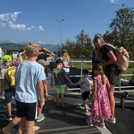 Eine Gruppe von Kindern und Erwachsenen geht auf einer Metallgitterfläche, wahrscheinlich in einem Park. Einige tragen Hüte und Turnschuhe. Eine Frau trägt eine Tasche. Bäume und Berge sind im Hintergrund.