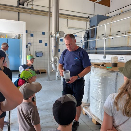 Ein Mann in einem blauen T-Shirt hält einen Becher mit Flüssigkeit und erklärt etwas einer Gruppe von Kindern in einer Fabrikumgebung.