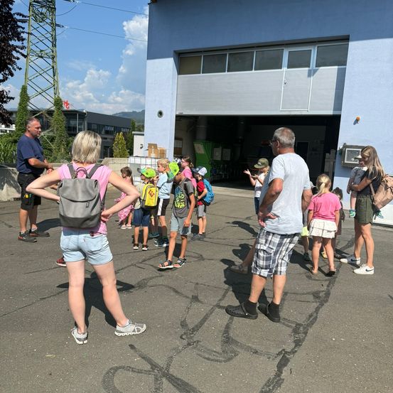 Eine Gruppe von Kindern und Erwachsenen versammelt sich vor einem Gebäude, möglicherweise einer Schule, mit einem Mann in einem blauen Shirt und einer Frau in einem rosa Shirt, die sie führen. Die Kinder tragen Rucksäcke und Turnschuhe, und einige halten sich an den Erwachsenen fest. Das Gebäude hat eine Garagentür und eine Wand mit einem Fenster.