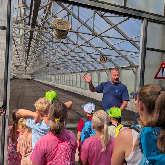 Ein Mann in einem blauen T-Shirt führt eine Gruppe von Kindern durch ein Gewächshaus. Die Kinder tragen Hüte und Rucksäcke.