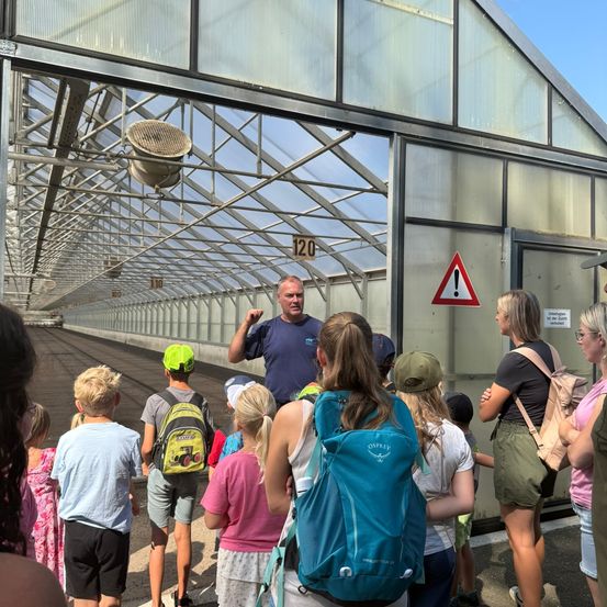 Eine Gruppe von Kindern und Erwachsenen steht in einem Gewächshaus, wobei ein Mann zu ihnen spricht. Das Gewächshaus hat einen Metallrahmen und Glasscheiben, und ein Warnhinweis ist an der Tür angebracht.