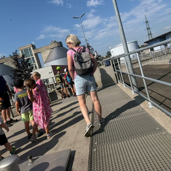 Eine Gruppe von Menschen, darunter eine Frau mit Rucksack, geht auf einem Betonweg mit Metallgittern. Sie sind in der Nähe eines Gebäudes mit einer kuppelförmigen Struktur und einer Straßenlaterne. Der Himmel ist blau mit einigen Wolken.