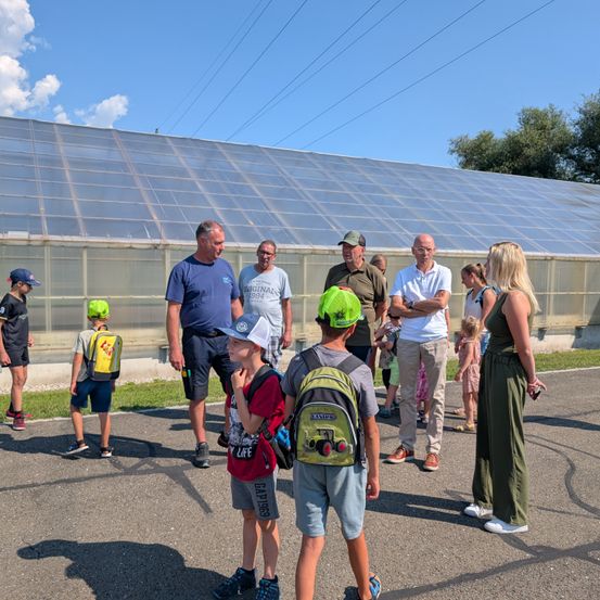 Eine Gruppe von Erwachsenen und Kindern steht vor einem großen Gewächshaus, wahrscheinlich auf einer Bauernhoftour. Die Kinder tragen Hüte und Rucksäcke.