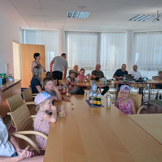 Eine Gruppe von Menschen, einschließlich Kindern, sitzt um einen langen Tisch in einem Raum mit großen Fenstern. Flaschen, Gläser und andere Gegenstände stehen auf dem Tisch. Einige Menschen stehen in der Nähe der Tür.