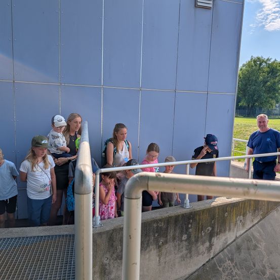 Eine Gruppe von Kindern und Erwachsenen steht auf einer Betonplattform mit Metallgeländern, möglicherweise auf einer Tour. Sie schauen auf etwas rechts. Ein Mann in blauer Uniform steht rechts.