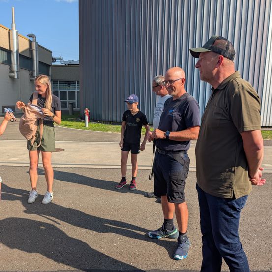Eine Gruppe von Menschen steht draußen in der Nähe eines Gebäudes, eine Person hält eine braune Tasche und eine andere eine Flasche.