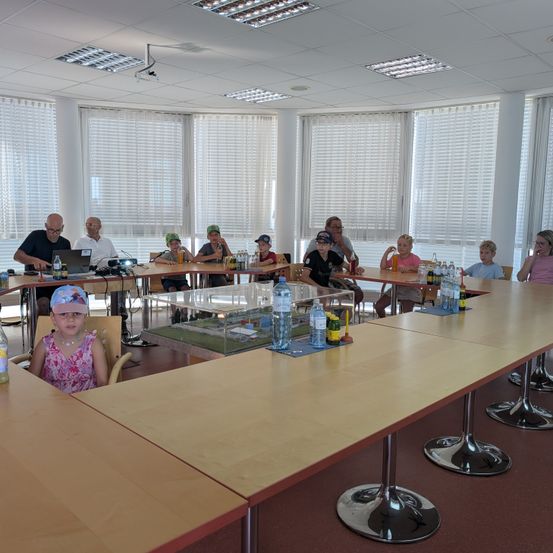 Eine Gruppe von Menschen, einschließlich Kindern, sitzt um einen langen Tisch in einem Konferenzraum mit großen Fenstern und einer Glasvitrine. Flaschen und Becher stehen auf dem Tisch.