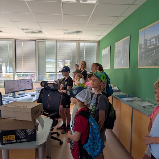 Eine Gruppe von Kindern in einem Klassenzimmer, einige mit Mützen und Rucksäcken, betrachten einen Computerbildschirm auf einem Schreibtisch.