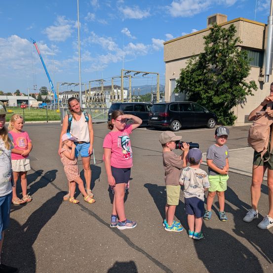 Eine Gruppe von Kindern und Erwachsenen steht auf einem Parkplatz, einige halten Kameras, mit einem Gebäude und Bäumen im Hintergrund.