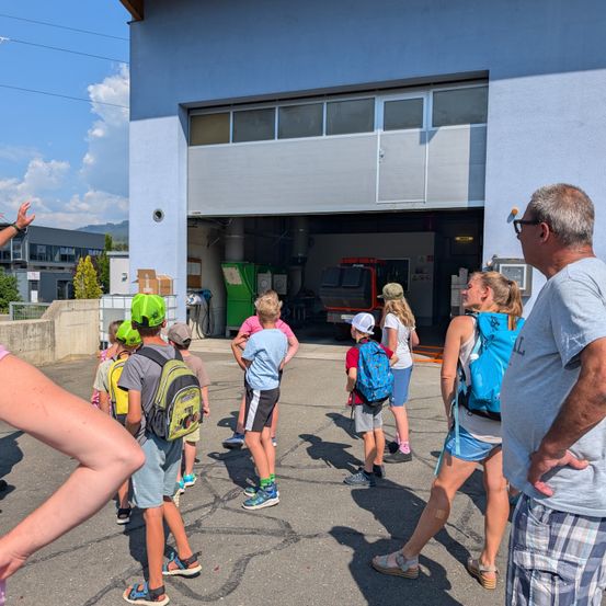 Eine Gruppe von Kindern und Erwachsenen steht vor einem Gebäude mit einem Feuerwehrauto im Inneren. Die Kinder tragen Rucksäcke und einige tragen Hüte. Die Erwachsenen stehen neben den Kindern.