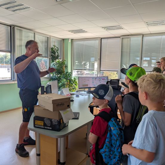 Ein Mann in einem blauen Shirt hält eine Präsentation für eine Gruppe von Kindern in einem Klassenzimmer. Die Kinder tragen Mützen und Rucksäcke. Es gibt einen Tisch mit Kisten und einem Computermonitor.