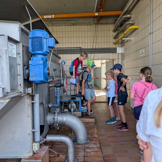 Eine Gruppe von Kindern, einige mit Mützen, stehen in einer Fabrik und betrachten eine große blaue Maschine mit Rohren und Ventilen.