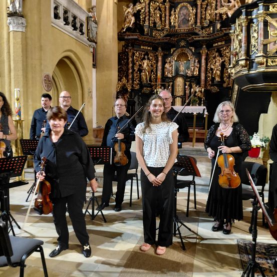 Eine Gruppe von Musikern, darunter ein junges Mädchen, steht in einer Kirche mit Violinen und Notenständern und lächelt für ein Foto.
