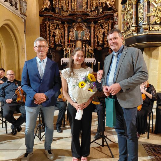 Ein junges Mädchen mit Blumen steht zwischen zwei Männern in einer Kirche, mit Musikern im Hintergrund.