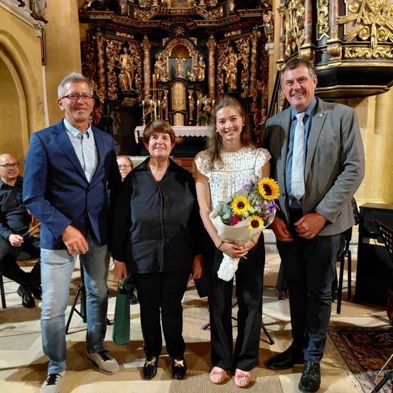 Vier Erwachsene und ein junges Mädchen stehen lächelnd vor einem Kirchenaltar und halten einen Blumenstrauß. Der Mann links trägt einen blauen Anzug, der Mann rechts einen grauen Anzug. Die Frau in der Mitte trägt eine schwarze Jacke.