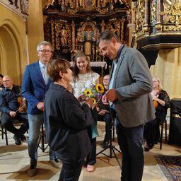 Ein Mann und eine Frau tauschen Blumen in einer Kirche aus, während Musiker im Hintergrund spielen.