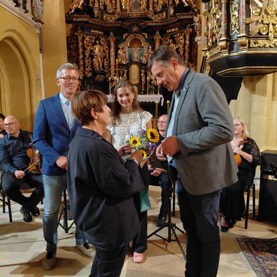 Ein Mann und eine Frau tauschen Blumen in einer Kirche aus, während Musiker im Hintergrund spielen.