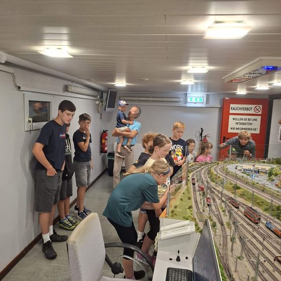 Eine Gruppe von Kindern und Erwachsenen versammelt sich um eine Modelleisenbahn, wobei eine Person die Steuerung bedient. Der Raum hat weiße Wände und eine Decke mit Lichtern.