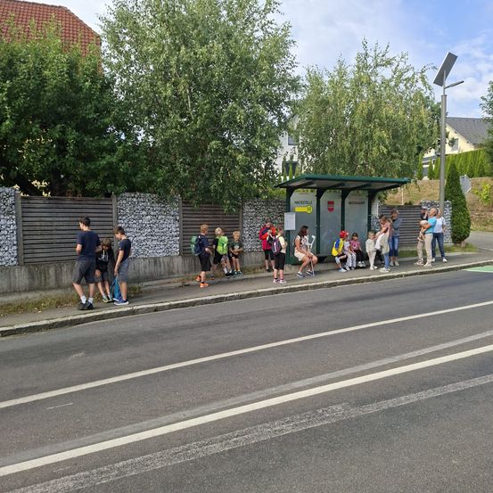 Eine Gruppe von Kindern und Erwachsenen versammelt sich an einer Bushaltestelle an einem sonnigen Tag. Einige sitzen auf der Bank, andere stehen. Im Hintergrund sind Bäume und Häuser zu sehen.