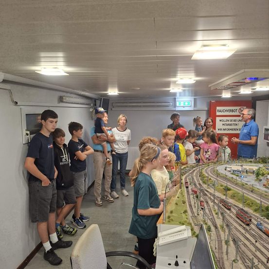 Eine Gruppe von Kindern und Erwachsenen versammelt sich um eine Miniatur-Eisenbahn, wobei ein Mann in einem blauen Hemd eine Präsentation hält. Der Raum hat eine Decke mit Lichtern und ein Schild, das Rauchverbot sagt.