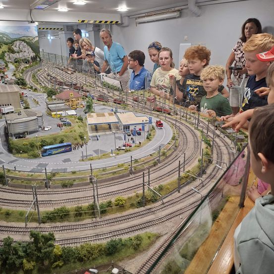 Eine Gruppe von Kindern und Erwachsenen versammelt sich um eine große Modelleisenbahn, um die feinen Details und Miniaturzüge auf den Gleisen zu beobachten.