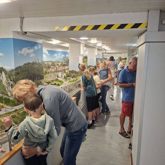 Eine Gruppe von Menschen, darunter ein Mann und ein Kind, betrachten eine Miniatur-Zugausstellung. Die Wände sind mit landschaftlichen Wandgemälden verziert.