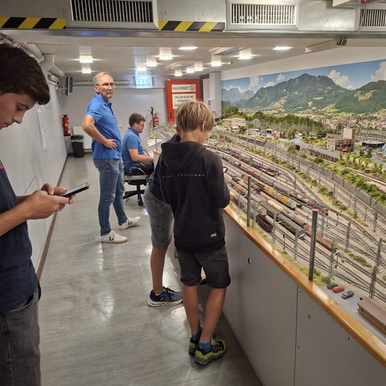 Vier Personen befinden sich in einem Raum mit einer Modelleisenbahn. Drei schauen auf die Züge, während ein Mann sein Handy benutzt.