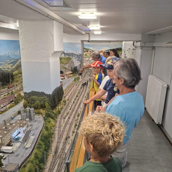 Eine Gruppe von Menschen, einschließlich Kindern, betrachten eine Modelleisenbahn mit einer landschaftlichen Kulisse. Die Anlage umfasst einen Bahnhof, Gleise und Miniaturgebäude.