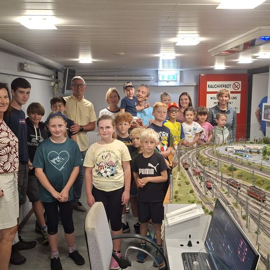 Eine Gruppe von Kindern und Erwachsenen steht in einem Raum mit einer Modelleisenbahn und lächelt für ein Foto. Ein Schild an der Wand zeigt 'Rauchverbot' und 'WOLL MITTEN UNS'.