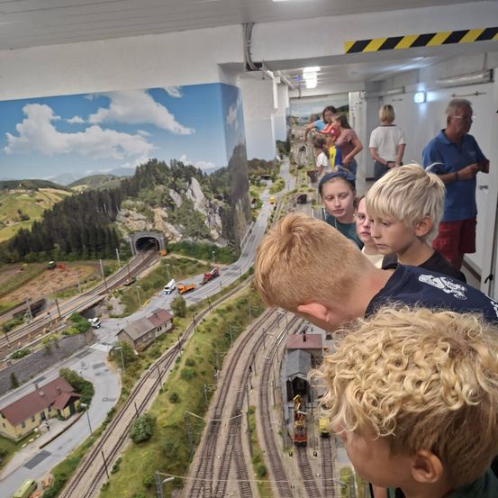 Eine Gruppe von Kindern und Erwachsenen betrachtet eine Modelleisenbahn mit einer malerischen Kulisse von Bergen und einem Tunnel.